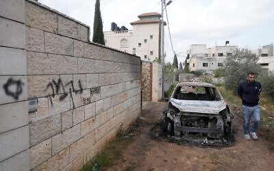 Un Palestinien examine un véhicule incendié à côté d'un graffiti en hébreu sur un mur où l'on peut lire « vengeance », à la suite d'une attaque perpétrée par des pro-implantations dans les villages voisins de la région de Naplouse, dans le village de Deir al-Hatab, en Cisjordanie, le 23 mars 2026. (Crédit : AP/Majdi Mohammed)