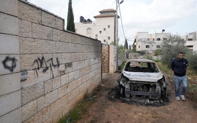Un Palestinien examine un véhicule incendié à côté d'un graffiti en hébreu sur un mur où l'on peut lire « vengeance », à la suite d'une attaque perpétrée par des pro-implantations dans les villages voisins de la région de Naplouse, dans le village de Deir al-Hatab, en Cisjordanie, le 23 mars 2026. (Crédit : AP/Majdi Mohammed)