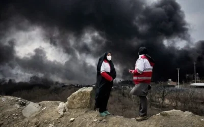 Deux femmes travaillant pour le Croissant-Rouge iranien se tiennent debout alors qu'un épais nuage de fumée consécutif à une frappe américano-israélienne contre des installations de stockage de pétrole, samedi soir, s'élève dans le ciel à Téhéran, en Iran, le 8 mars 2026. (Photo AP/Vahid Salemi, Dossier)
