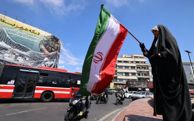 Une femme agite le drapeau national iranien devant un panneau d'affichage géant sur lequel on peut lire « Le détroit d'Ormuz reste fermé », sur la place de la Révolution à Téhéran, le 12 avril 2026. (Crédit: Atta Kenare/AFP)