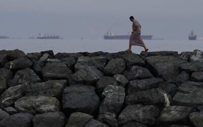 Un homme marchant le long du rivage tandis que des pétroliers et des cargos s’alignent dans le détroit d’Ormuz, vu depuis Khor Fakkan, aux Émirats arabes unis, le 11 mars 2026. (Crédit : Altaf Qadri/AP Photo)