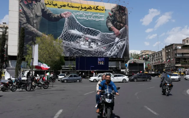 Des véhicules et des motos passent devant un panneau anti-américain représentant un avion américain entrant dans un filet de pêche des forces armées iraniennes, avec des inscriptions en farsi : « Le détroit d’Ormuz restera fermé, tout le golfe Persique est notre terrain de chasse », sur la place de la Révolution islamique, dans le centre-ville de Téhéran, en Iran, le 5 avril 2026. (Crédit: AP Photo/Vahid Salemi)