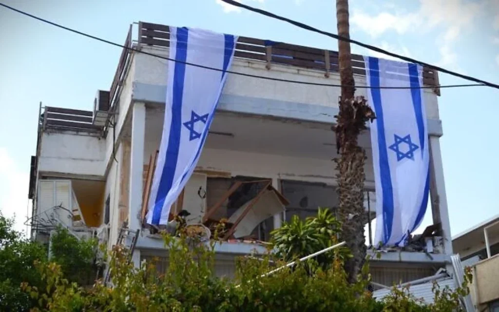 Des drapeaux israéliens ornent la façade d'un bâtiment endommagé par un missile à fragmentation iranien, le 5 avril 2026. (Stav Levaton/Times of Israel) Des drapeaux israéliens ornent la façade d'un bâtiment endommagé par un missile à fragmentation iranien, le 5 avril 2026. (Stav Levaton/Times of Israel)