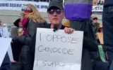 Le leader de Massive Attack, Robert Del Naja, tient une pancarte sur laquelle on peut lire « Je m'oppose au génocide, je soutiens Palestine Action », le 11 avril 2026, en solidarité avec le groupe anti-israélien interdit car répertorié comme organisation terroriste après avoir pénétré dans des usines de défense et une base de la Royal Air Force en faisant des millions de livres de dégâts et en agressant un policier. (Capture d'écran via La Jornada sur YouTube)