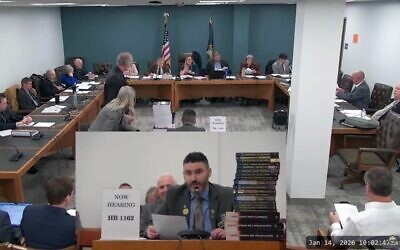 Matt Sabourin dit Choinière (au centre), député républicain de l'État du New Hampshire, témoigne devant une commission d'État au sujet de son projet d'amendement visant à nommer un négationniste au sein de la commission d'État chargée de l'éducation sur la Shoah, le 14 janvier 2026. Il est entouré d'une pile d'ouvrages négationnistes. (Capture d'écran via JTA)