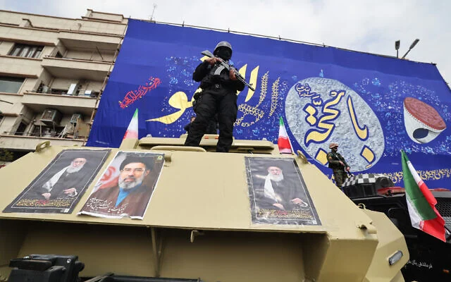 Un membre des forces de sécurité iraniennes monte la garde sur un véhicule blindé recouvert de photos, de gauche à droite, du dirigeant suprême iranien assassiné Ali Khamenei, de son fils et successeur Mojtaba Khamenei et du guide suprême fondateur Ruhollah Khomeini, lors du cortège funèbre des commandants des Gardiens de la Révolution tués lors de la guerre, le 11 mars 2026. (Crédit : Atta Kenare/AFP)