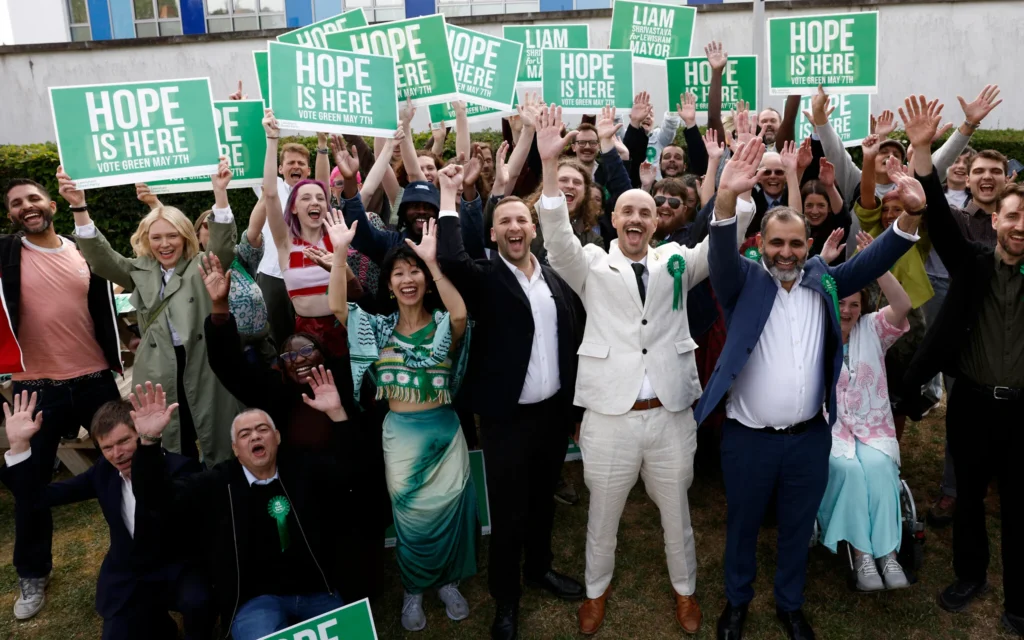 Le chef du Parti vert, Zack Polanski (au centre), célébrant avec le maire de Lewisham nouvellement élu par son parti, Liam Shrivastava (au centre à droite), et des partisans, devant le centre de dépouillement de Lewisham, au sud de Londres, le 8 mai 2026. (Crédit : Brook Mitchell/AFP)