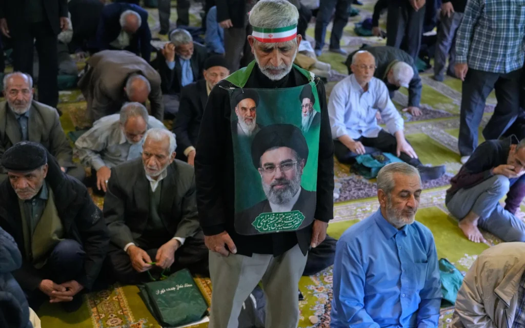 Un fidèle iranien porte une banderole sur laquelle figurent les portraits du Guide suprême, l’ayatollah Mojtaba Khamenei (en bas), du défunt Guide suprême, l’ayatollah Ali Khamenei (en haut à droite), et du défunt fondateur de la révolution, l’ayatollah Khomeini, lors de la cérémonie de la prière du vendredi sur le campus de l’université de Téhéran, à Téhéran, en Iran, le 24 avril 2026. (Crédit : Vahid Salemi/AP)