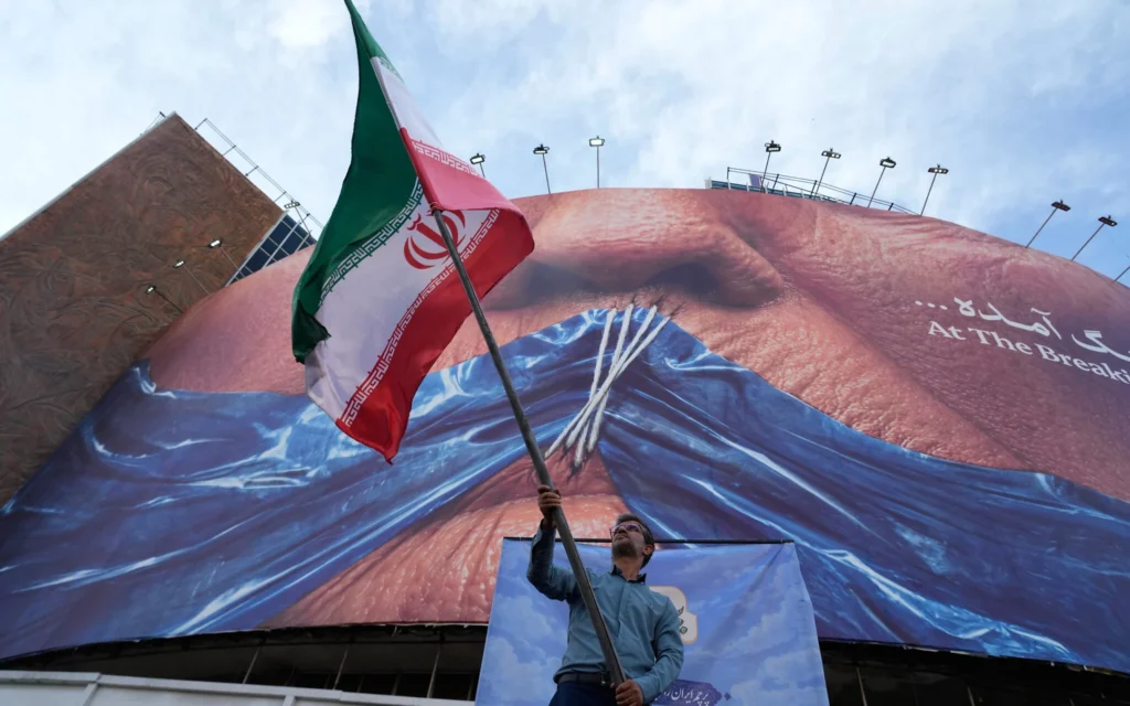 Un homme brandit un drapeau iranien dans le cadre d'une campagne pro-gouvernementale, sous un panneau d'affichage représentant le détroit d'Ormuz et le président américain Donald Trump aux lèvres cousues, sur une place du centre-ville de Téhéran, le 6 mai 2026. (Crédit:  AP/Vahid Salemi)