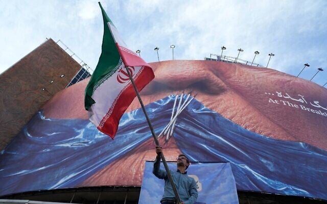 Un homme agitant un drapeau iranien dans le cadre d’une campagne pro-régime sous un panneau d’affichage représentant le détroit d’Ormuz et les lèvres cousues du président américain Donald Trump, sur une place du centre-ville de Téhéran, le 6 mai 2026. (Crédit : Vahid Salemi/AP)