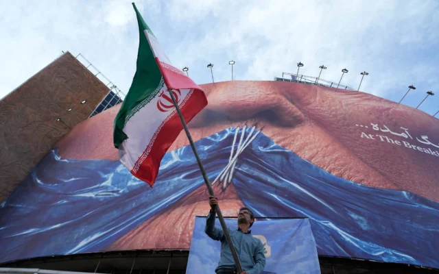 Un homme agite un drapeau iranien dans le cadre d'une campagne pro-gouvernementale, sous un panneau d'affichage représentant le détroit d'Ormuz et le président américain Donald Trump aux lèvres cousues, sur une place du centre-ville de Téhéran, le 6 mai 2026. (Crédit: AP Photo/Vahid Salemi)