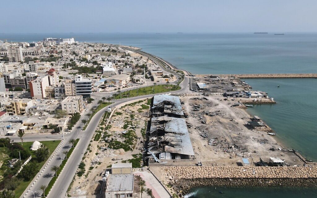 En arrière-plan des navires dans le détroit d'Ormuz, on aperçoit les dégâts causés, selon des témoins, par plusieurs frappes aériennes, sur un quai de pêche du port de l'île de Qeshm, en Iran, le 13 avril 2026. (Crédit : Asghar Besharati/AP)