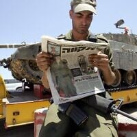 Un soldat israélien lisant un journal sur lequel figure une photo de Gilad Shalit, tandis qu'un char se prépare à se déployer près de la frontière avec Gaza, le 26 juin 2006. (Crédit : Menahem Kahana/AFP)