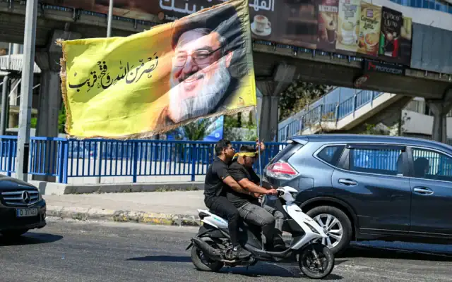 Des hommes circulent en scooter en arborant une banderole à l'effigie de Hassan Nasrallah, le chef assassiné du groupe terroriste chiite libanais Hezbollah, dans la banlieue sud de Beyrouth, le 26 juin 2026, au plus fort de l'Achoura, une période de dix jours commémorant l'assassinat, au VIIe siècle, de l'imam Hussein, petit-fils du prophète Mahomet. (Crédit: Joseph Eid/AFP)