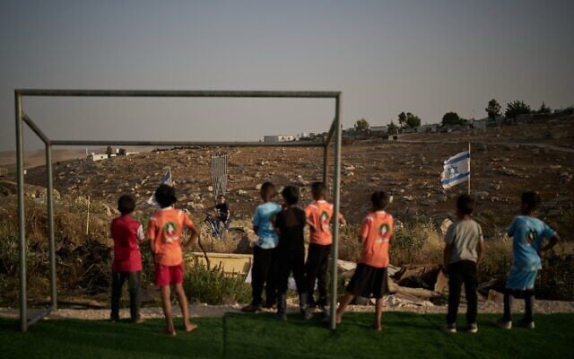 De jeunes garçons Palestiniens regardent un jeune résident d'implantation israélien après que leur ballon de football, tombé au-delà d'une clôture barbelée entourant un petit terrain près de l'implantation de Carmel, leur a été confisqué, dans le village bédouin d'Umm al-Khair, en Cisjordanie, le 12 mai 2026. (Crédit : AP Photo/Leo Correa)