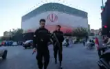 Des policiers marchant devant une immense banderole arborant le drapeau de la République islamique, sur la place de la Révolution islamique, à Téhéran, en Iran, le 14 juin 2026. (Crédit : Vahid Salemi/AP)