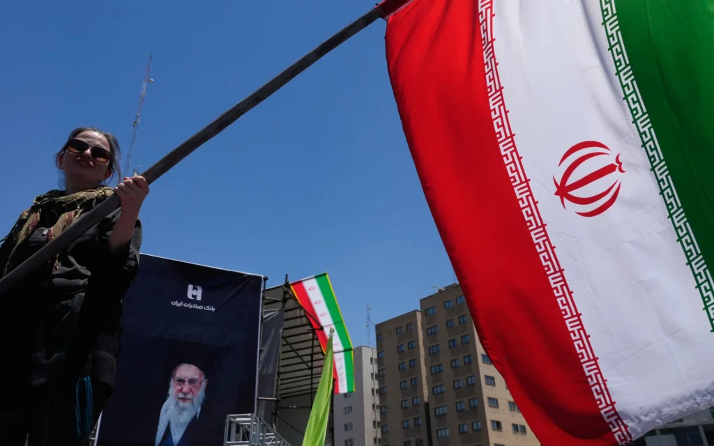 Une femme agitant un drapeau pré-Révolution islamique lors d'une campagne pro-régime, un portrait du guide suprême assassiné, l'ayatollah Ali Khamenei, éliminé lors de frappes américaines et israéliennes le 28 février, exposé à l'arrière-plan, dans le centre-ville de Téhéran, en Iran, le 15 juin 2026. (Crédit : Vahid Salemi/AP Photo)