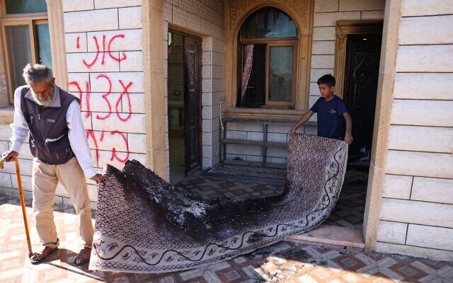 Un Palestinien et un enfant sortant un tapis brûlé d'une mosquée dont l'entrée a été incendiée et les murs recouverts de graffitis en hébreu, lors d'une attaque qui aurait été perpétrée par des résidents d'implantations, dans un village palestinien près de Hébron, en Cisjordanie, le 19 juillet 2026. (Crédit : Hazem Bader/AFP)