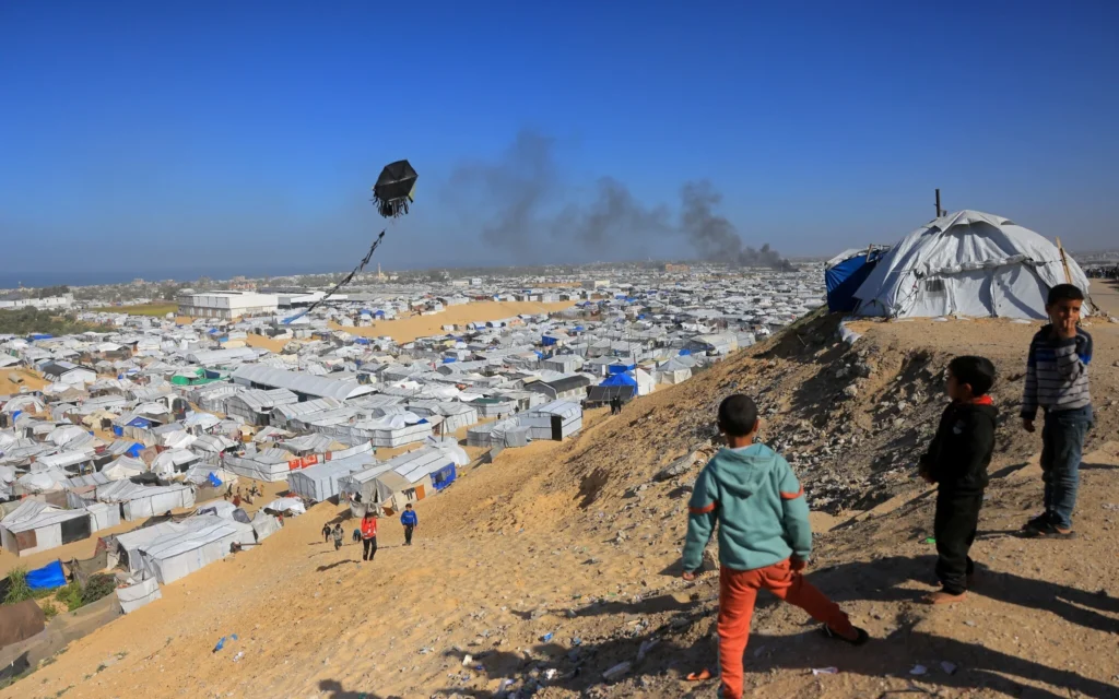 Des jeunes garçons font voler un cerf-volant avec, en arrière-plan, la fumée s'élevant du centre d'accueil de Gath, qui héberge des Palestiniens déplacés, après une frappe aérienne israélienne à l'ouest de Khan Younis, dans le sud de la bande de Gaza, le 31 janvier 2026. (Crédit: Bashar Taleb/AFP)