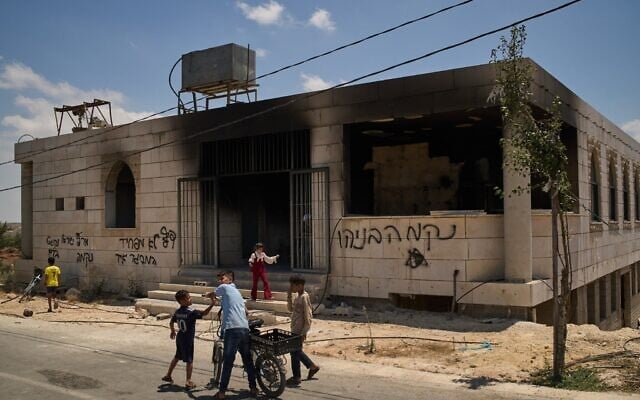 Des enfants se tiennent devant une mosquée incendiée lors d'une attaque menée par des résidents d'implantations israéliens tôt ce matin dans le village de Qusra, en Cisjordanie, le 26 juillet 2026. Sur le mur de l'édifice, un graffiti en hébreu indique « Vengeance Benayahu », en référence à un résident d'implantations israélien tué vendredi dernier lors d'affrontements avec des Palestiniens dans le village de Tell. (Crédit : AP Photo/Leo Correa)