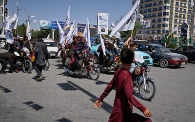 Un garçon passant devant des membres des talibans à moto qui agitent des drapeaux lors des célébrations marquant le cinquième anniversaire du retrait américain d'Afghanistan et du retour au pouvoir des talibans, à Kaboul, en Afghanistan, le 15 août 2026. (Crédit : Nava Jamshidi/AP)