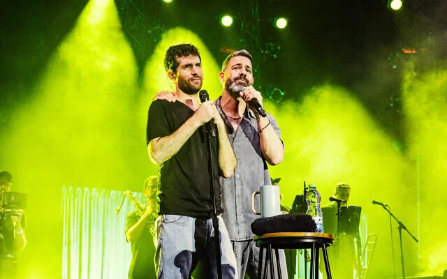 L'ancien otage Sagui Dekel-Chen et Omri Glickman, chanteur principal du groupe HaTivka6, interprétant une chanson composée par Dekel-Chen pendant sa captivité lors d'un spectacle sur la pelouse du kibboutz Nir Oz dans le cadre du Festival d'Israël, le 10 août 2026. (Crédit : Orit Pnini)