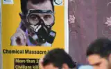 Des personnes sont assises face à une affiche représentant le président syrien Bachar al-Assad portant un masque à gaz, lors d'un événement organisé le 20 août 2023 dans la ville d'Afrin, au nord du pays, contrôlée par les rebelles, pour marquer le 10e anniversaire des attaques chimiques à  la Ghouta, près de la capitale. (Crédit : Rami al Sayed/AFP)
