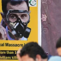 Des personnes sont assises face à une affiche représentant le président syrien Bachar al-Assad portant un masque à gaz, lors d'un événement organisé le 20 août 2023 dans la ville d'Afrin, au nord du pays, contrôlée par les rebelles, pour marquer le 10e anniversaire des attaques chimiques à  la Ghouta, près de la capitale. (Crédit : Rami al Sayed/AFP)