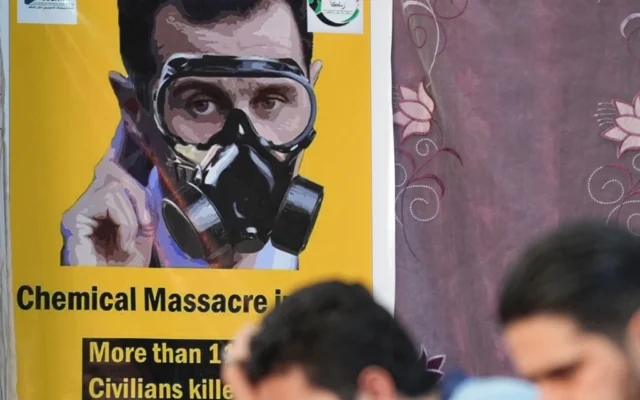 Des personnes sont assises face à une affiche représentant le président syrien Bachar al-Assad portant un masque à gaz, lors d'un événement organisé le 20 août 2023 dans la ville d'Afrin, au nord du pays, contrôlée par les rebelles, pour marquer le 10e anniversaire des attaques chimiques à  la Ghouta, près de la capitale. (Crédit: Rami al Sayed/AFP)