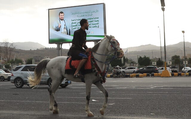 Un homme passe à cheval sous un panneau d'affichage montrant le chef houthi Abdul Malik al-Houthi accompagné d'une phrase du Coran, qui se lit en arabe : « C'est par la grâce d'Allah et par Sa miséricorde qu'ils se réjouissent ; c'est mieux que tout ce qu'ils accumulent », à Sanaa, au Yémen, le dimanche 6 septembre 2026. (Crédit :  AP/Osamah Abdulrahman)