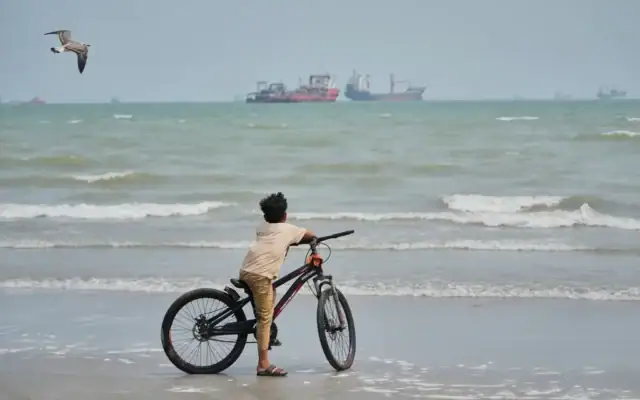 Une mouette vole au-dessus d’un garçon debout près de son vélo sur une plage surplombant des navires commerciaux ancrés dans le détroit d’Ormuz, au large de Bandar Abbas, en Iran, le 7 septembre 2026. (Crédit : Vahid Salemi/AP)