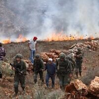 Des soldats israéliens marchent aux côtés d'un agriculteur palestinien tandis que d'autres tentent d'éteindre les incendies qui ravagent leurs terres, après que des soldats israéliens ont tiré des gaz lacrymogènes sur eux alors qu'ils récoltaient leurs olives, provoquant ainsi l'embrasement des champs asséchés, dans le village d'Asira al-Qibliya, au sud de la ville de Naplouse, le 3 octobre 2026. (Crédit : Jaafar Ashtiyeh/AFP)