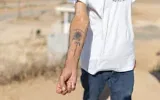 Danny, un habitant du kibboutz Nahal Oz, montrant le tatouage à l'encre noire représentant un tournesol qu'il porte sur l'avant-bras, accompagné d'une inscription en hébreu signifiant « Le chemin vers l'espoir », qu'il s'est fait faire en mémoire du pogrom perpétré par le groupe terroriste palestinien du Hamas le 7 octobre 2023, à Nahal Oz, dans le sud d'Israël, le 22 octobre 2026. (Crédit : Ilia Yefimovich/AFP)