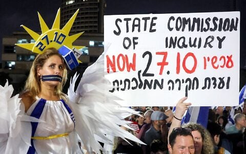 Une manifestante, la bouche recouverte d’une fermeture éclair et vêtue d’un costume, enveloppée dans le drapeau d'Israël, prenant la pose lors d’une manifestation anti-gouvernement, sur la Place des Otages, à Tel Aviv, le 24 octobre 2026, quelques jours avant les élections législatives. (Crédit : Jack Guez/AFP)