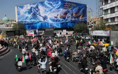 Des motards passent devant un panneau publicitaire représentant un héros légendaire persan et une silhouette volante frappant des porte-avions américains, accompagné d’un message en anglais et en farsi : "Une nouvelle puissance s’élève du golfe Persique", "L’armée de Satan sera noyée dans le golfe Persique", lors d’un défilé de motards organisé par l’État et baptisé "Janfada", ce qui signifie "Sacrifice pour l’Iran", à Téhéran, en Iran, le 25 septembre 2026. (Crédit: AP/Vahid Salemi)