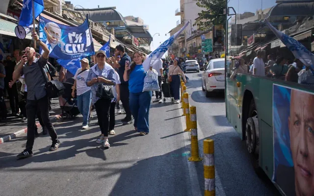 Des partisans du Premier ministre Benjamin Netanyahu défilent en brandissant des drapeaux du Likud, à côté d’un bus arborant une affiche électorale à l’effigie de son principal rival, Gadi Eisenkot, au marché Mahane Yehuda de Jérusalem, le 9 octobre 2026. (Crédit : Ohad Zwigenberg/AP)
