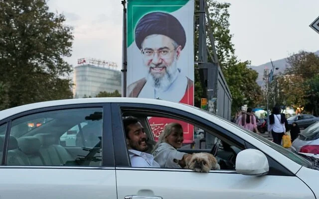 Un homme et une femme passant en voiture, accompagnés d’un chien, devant un portrait du guide suprême iranien Mojtaba Khamenei, à Téhéran, en Iran, le 21 octobre 2026. (Crédit : Vahid Salemi/AP) Un homme et une femme passant en voiture, accompagnés d’un chien, devant un portrait du guide suprême iranien Mojtaba Khamenei, à Téhéran, en Iran, le 21 octobre 2026. (Crédit : Vahid Salemi/AP)