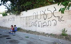 Children sit next to a wall in the Arab-Israeli village of Abu Ghosh on June 18, 2013 where anti-Arab graffiti was sprayed overnight. The graffiti reads “racism or assimilation” and “Arabs out.” (photo credit: Flash90).