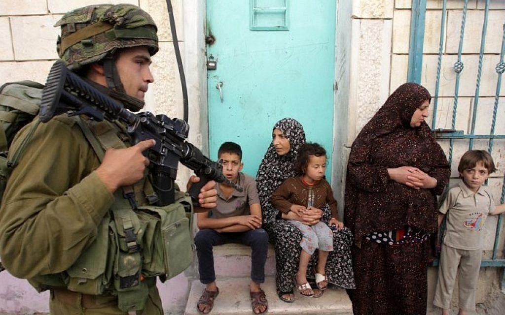 An Israeli soldier walks past Palestinians in the West Bank village of Tafoh, near Hebron on June 15, 2014, as Israeli army searches for three teenagers who went missing near a West Bank settlement. photo credit: AFP/HAZEM BADER) An Israeli soldier walks past Palestinians in the West Bank village of Tafoh, near Hebron on June 15, 2014, as Israeli army searches for three teenagers who went missing near a West Bank settlement. photo credit: AFP/HAZEM BADER)