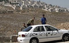 Palestinians stand next to one of ten cars that were sprayed with racist slogans that read in Hebrew "All the Arabs are enemies" in the east Jerusalem neighborhood of Beit Hanina, next to the Jewish settlement of Ramat Shlomo (background), on June 23, 2014. (photo credit: Ahmad Gharabli/AFP)