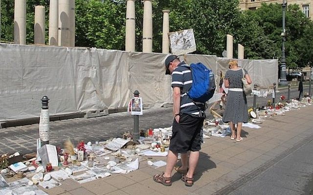 Passersby look at the Holocaust-related and other memorabilia left by citizens protesting the monument to the 1944 German occupation, under construction behind sheeting across the street in downtown Budapest. (Photo credit: JTA/Ruth Ellen Gruber)