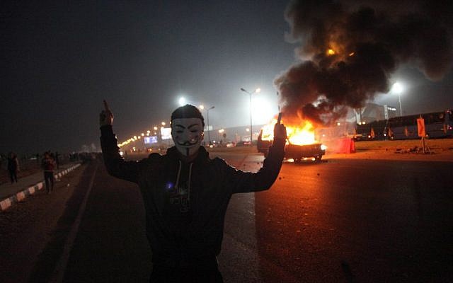 An Egyptian man wearing a mask of the Anonymous movement near a burning car outside a sports stadium in a Cairo's northeast district, on February 8, 2015, during clashes between supporters of Zamalek football club and security forces. (photo credit: AFP PHOTO / STR) An Egyptian man wearing a mask of the Anonymous movement near a burning car outside a sports stadium in a Cairo's northeast district, on February 8, 2015, during clashes between supporters of Zamalek football club and security forces. (photo credit: AFP PHOTO / STR)