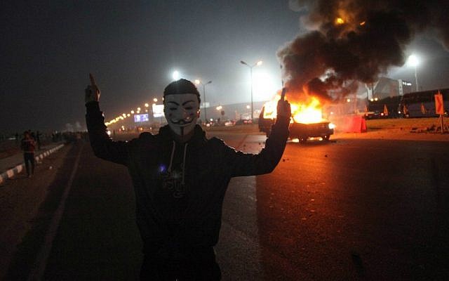 An Egyptian man wearing a mask of the anonymous movement gestures near a burning car outside a sports stadium in a Cairo's northeast district, on February 8, 2015 during clashes between supporters of Zamalek football club and security forces. (photo credit: AFP/STR) An Egyptian man wearing a mask of the anonymous movement gestures near a burning car outside a sports stadium in a Cairo's northeast district, on February 8, 2015 during clashes between supporters of Zamalek football club and security forces. (photo credit: AFP/STR)