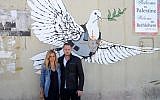 Journalist Mariana Van Zeller, left, and chef Michael Voltaggio, hosts of Travel Channel's "Breaking Borders," pose in front of an art piece showing a dove in a bulletproof vest by graffiti artist Banksy in Bethlehem. (Photo credit: AP Photo/Travel Channel)