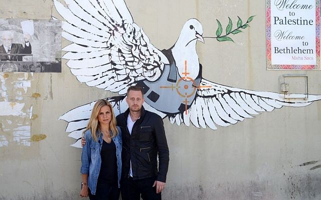 Journalist Mariana Van Zeller, left, and chef Michael Voltaggio, hosts of Travel Channel's "Breaking Borders," pose in front of an art piece showing a dove in a bulletproof vest by graffiti artist Banksy in Bethlehem. (Photo credit: AP Photo/Travel Channel) Journalist Mariana Van Zeller, left, and chef Michael Voltaggio, hosts of Travel Channel's "Breaking Borders," pose in front of an art piece showing a dove in a bulletproof vest by graffiti artist Banksy in Bethlehem. (Photo credit: AP Photo/Travel Channel)