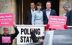 Protestors demonstrate as British Prime Minister and leader of the Conservative party David Cameron (R) leaves with his wife Samantha after voting at a polling station in Spelsbury on May 7, 2015 as Britain holds a general election (photo credit: AFP/ LEON NEAL)