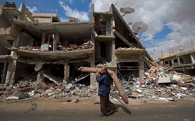 In this April 14, 2016 image, a Syrian man carries a carpet as walk through a devastated part of the town of Palmyra as families load their belongings onto a bus in the central Homs province, Syria. (AP Photo/Hassan Ammar)