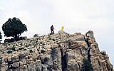 In this Saturday, May 9, 2015 photo, a Hezbollah fighter stands on a hill next to the group's yellow flag in the fields of the Syrian town of Assal al-Ward in the mountainous region of Qalamoun, Syria.  (AP Photo/Bassem Mroue, File)