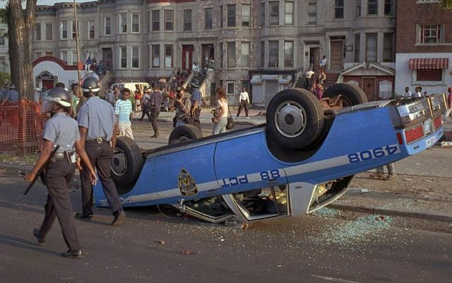 In this Aug. 21, 1991 file photo, New York City Police Department officers, in riot gear, walk past a police car that was overturned by rioters in the Crown Heights section of the Brooklyn borough of New York. (AP/David Burns) In this Aug. 21, 1991 file photo, New York City Police Department officers, in riot gear, walk past a police car that was overturned by rioters in the Crown Heights section of the Brooklyn borough of New York. (AP/David Burns)