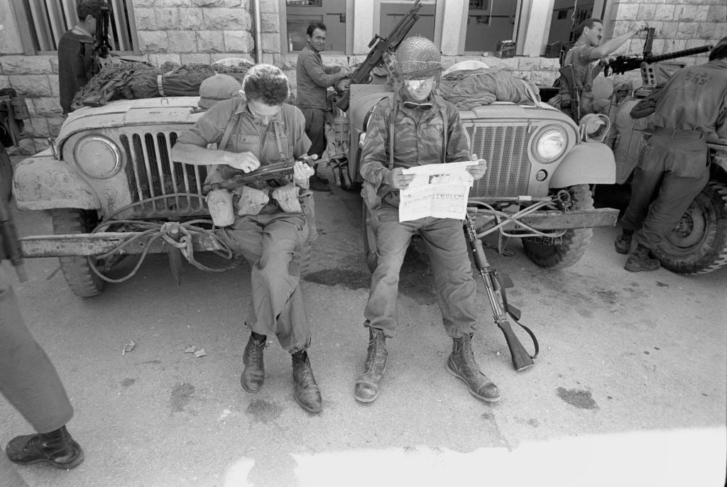 8 A soldier in the Paratroopers Brigade's reserve reconnaissance company cleans his rifle as his injured comrade reads the newspaper near the Western Wall on June 7, 1967. (Micha Bar-Am/Defense Ministry's IDF Archive)